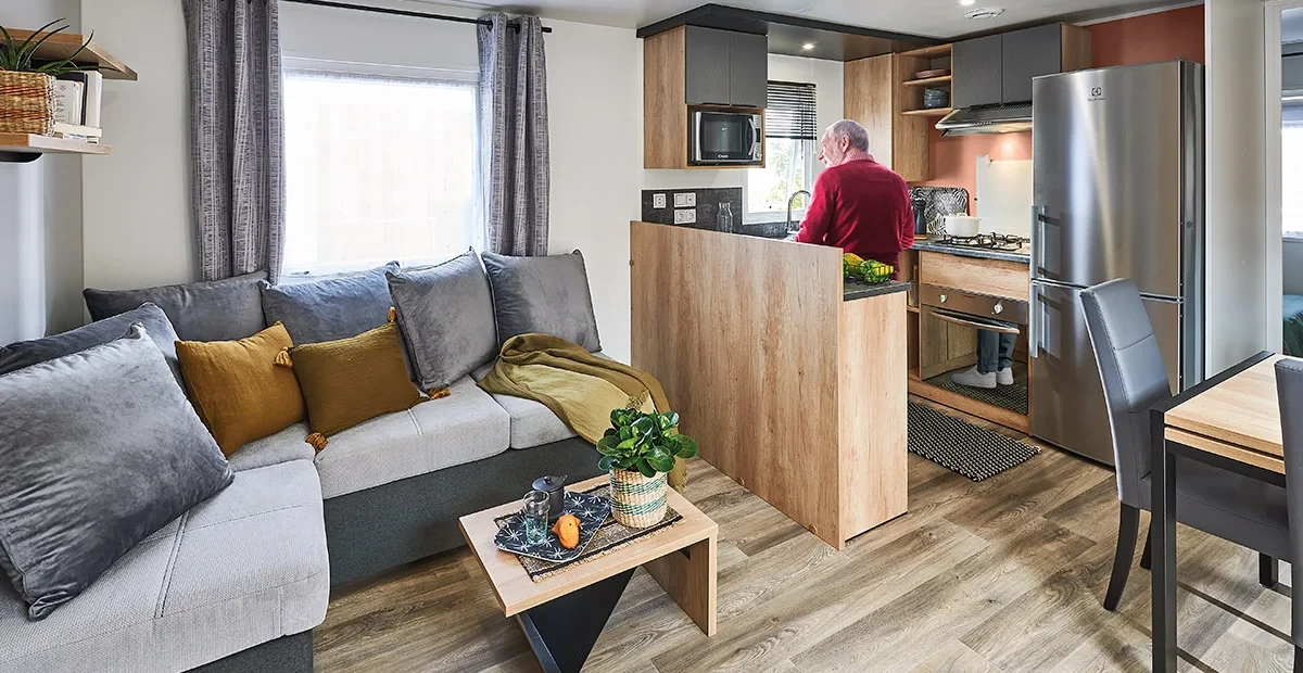 Séjour d’un mobil-home du Camping le ruisseau, avec canapé d’angle, table basse et cuisine ouverte où une personne prépare un repas.