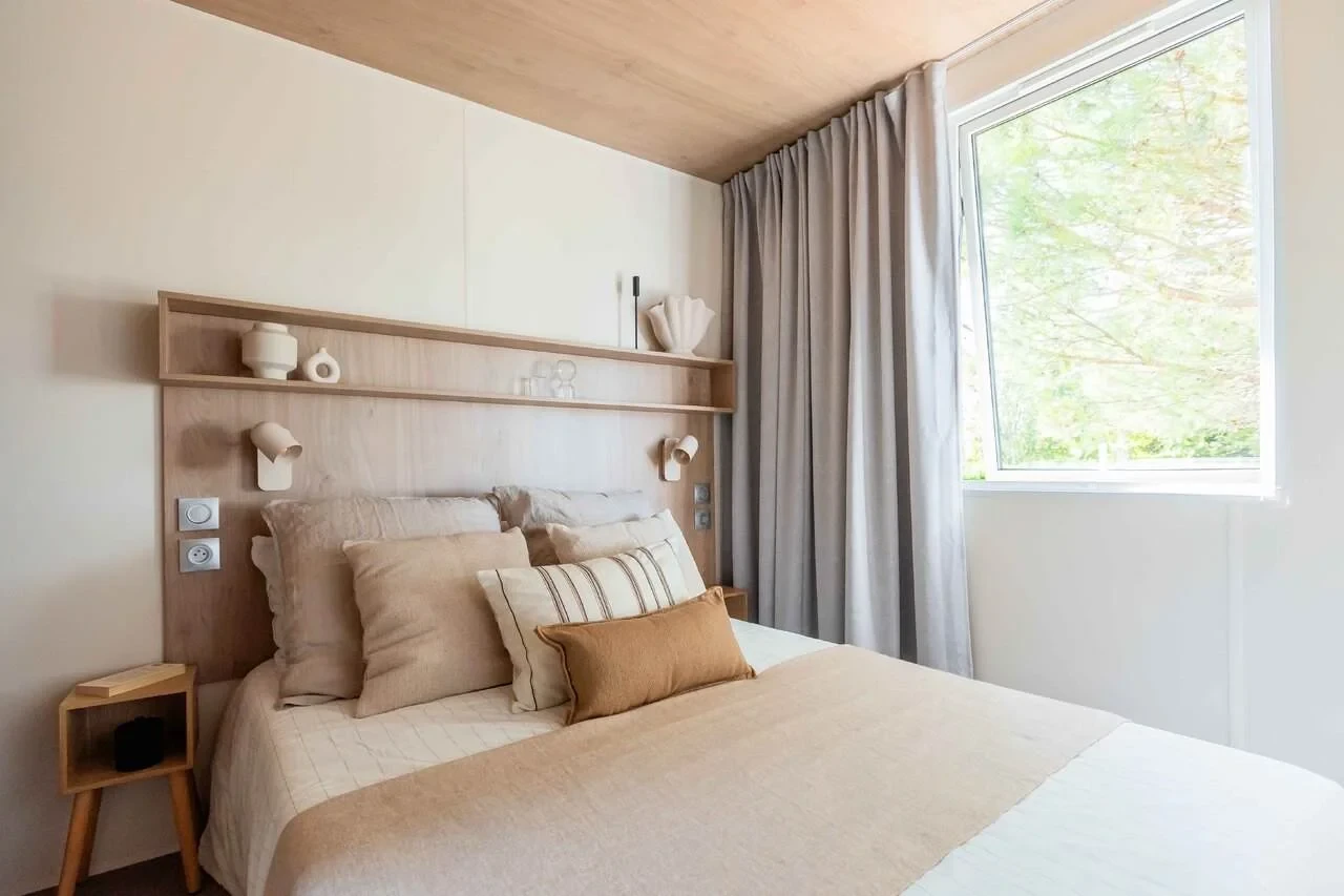 Lit double avec tête de lit en bois, liseuses et fenêtre bordée de rideaux gris.