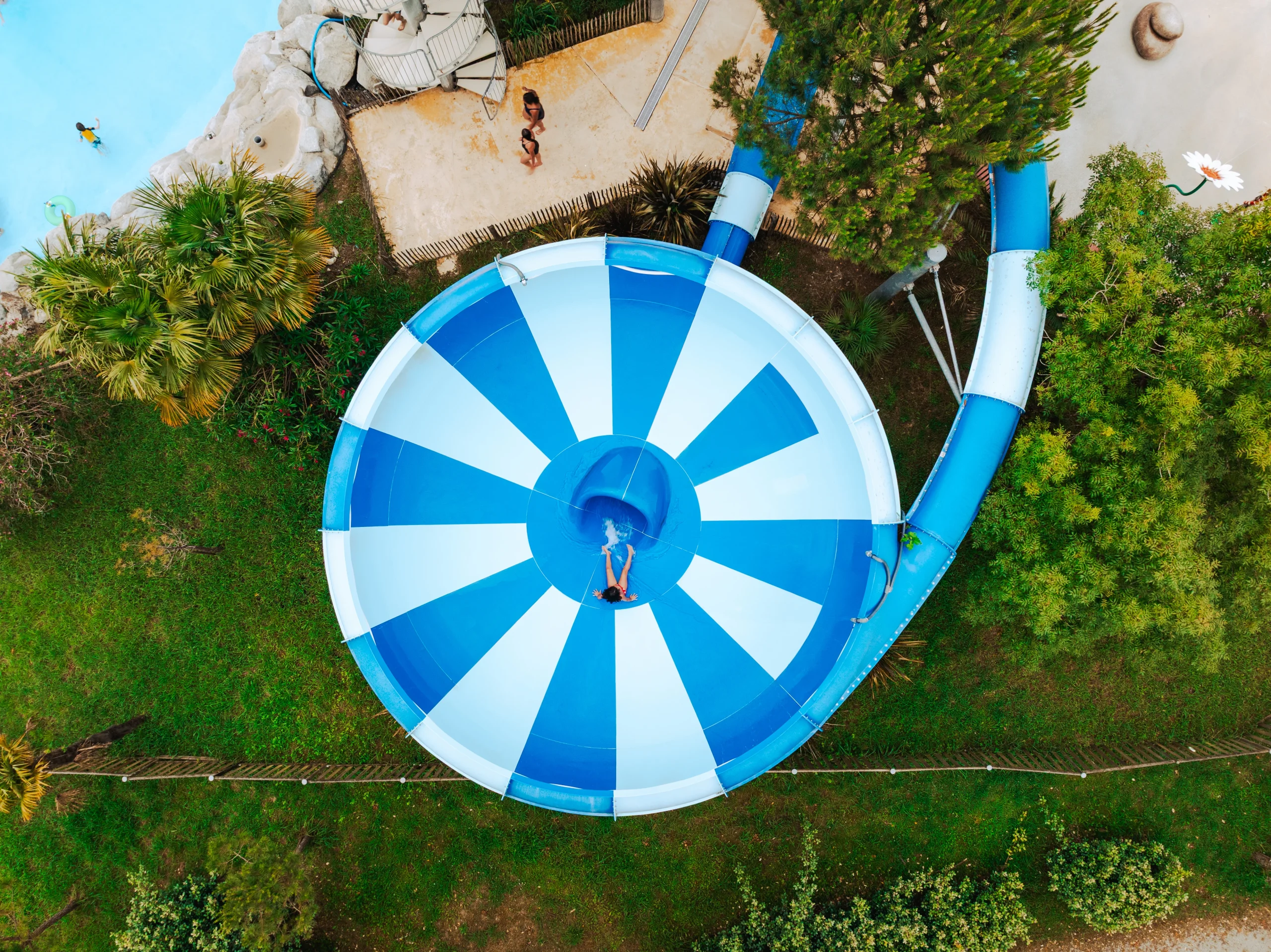 Vu du dessus, un baigneur glisse vers l’ouverture centrale du toboggan aquatique à entonnoir bleu et blanc du Camping le ruisseau, camping en bord de mer à Bidart.