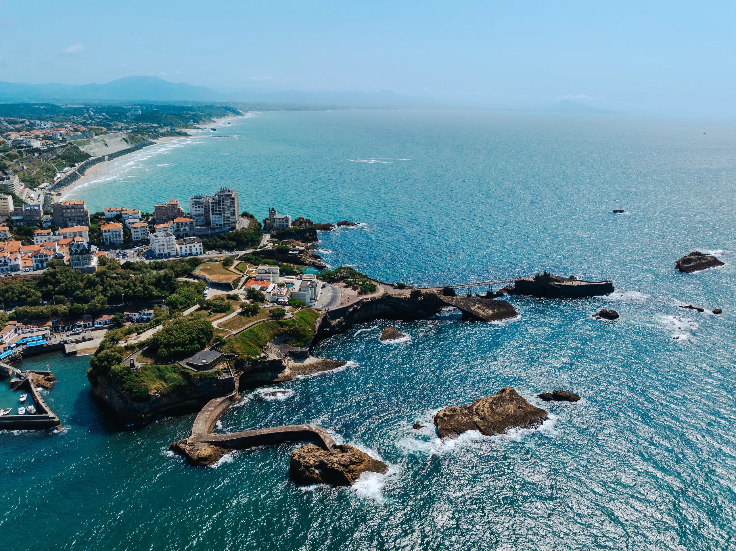Vue aérienne de la côte basque, avec un petit port, des falaises et une passerelle menant à un îlot rocheux.