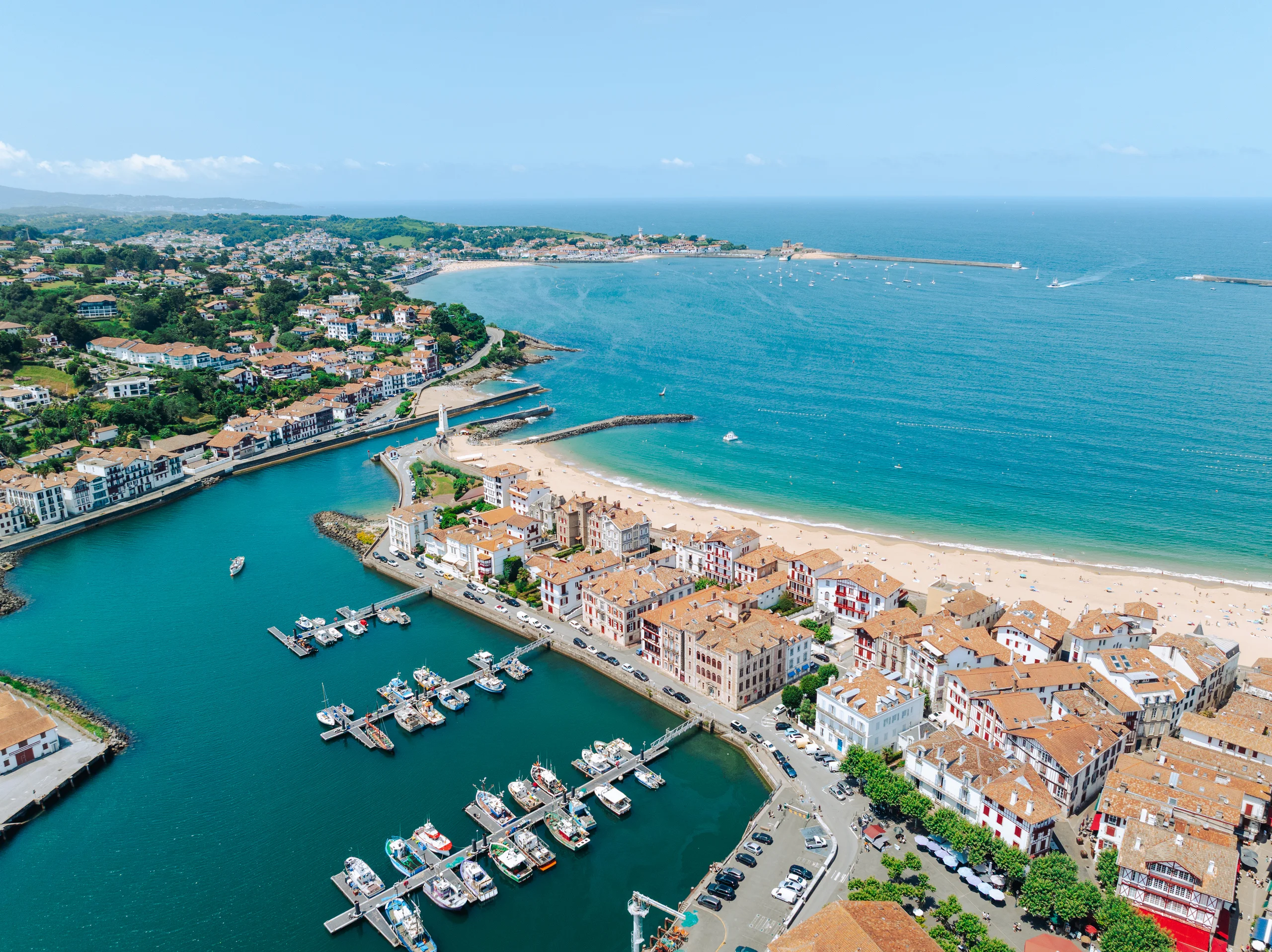 Vue aérienne d’un port de pêche et d’une grande baie, séparés par des maisons aux toits de tuiles et une plage de sable.