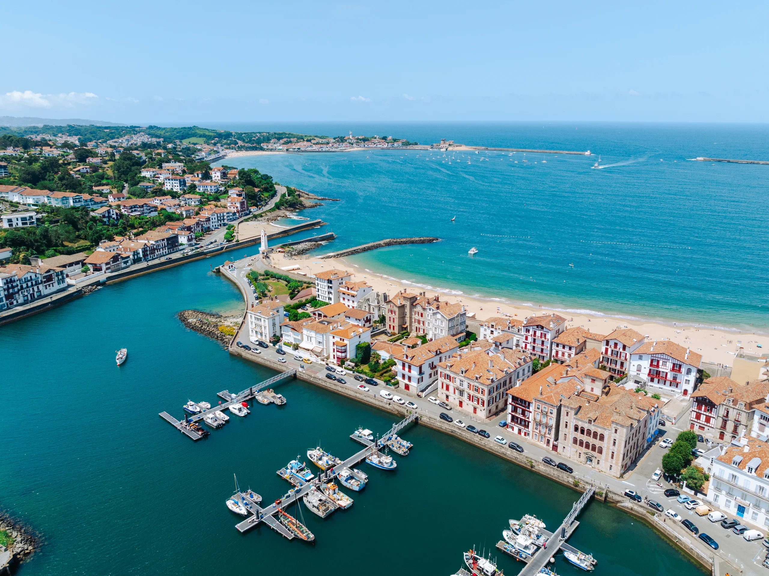 Vue aérienne d’un port bordé de maisons aux volets rouges, avec des bateaux à quai et une grande baie sablonneuse.