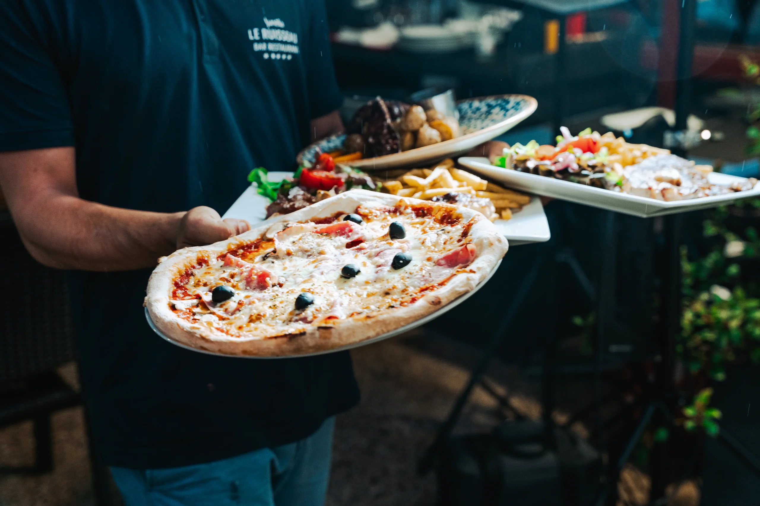 Une pizza et plusieurs assiettes sont portées en salle au Camping le ruisseau, camping à Bidart.