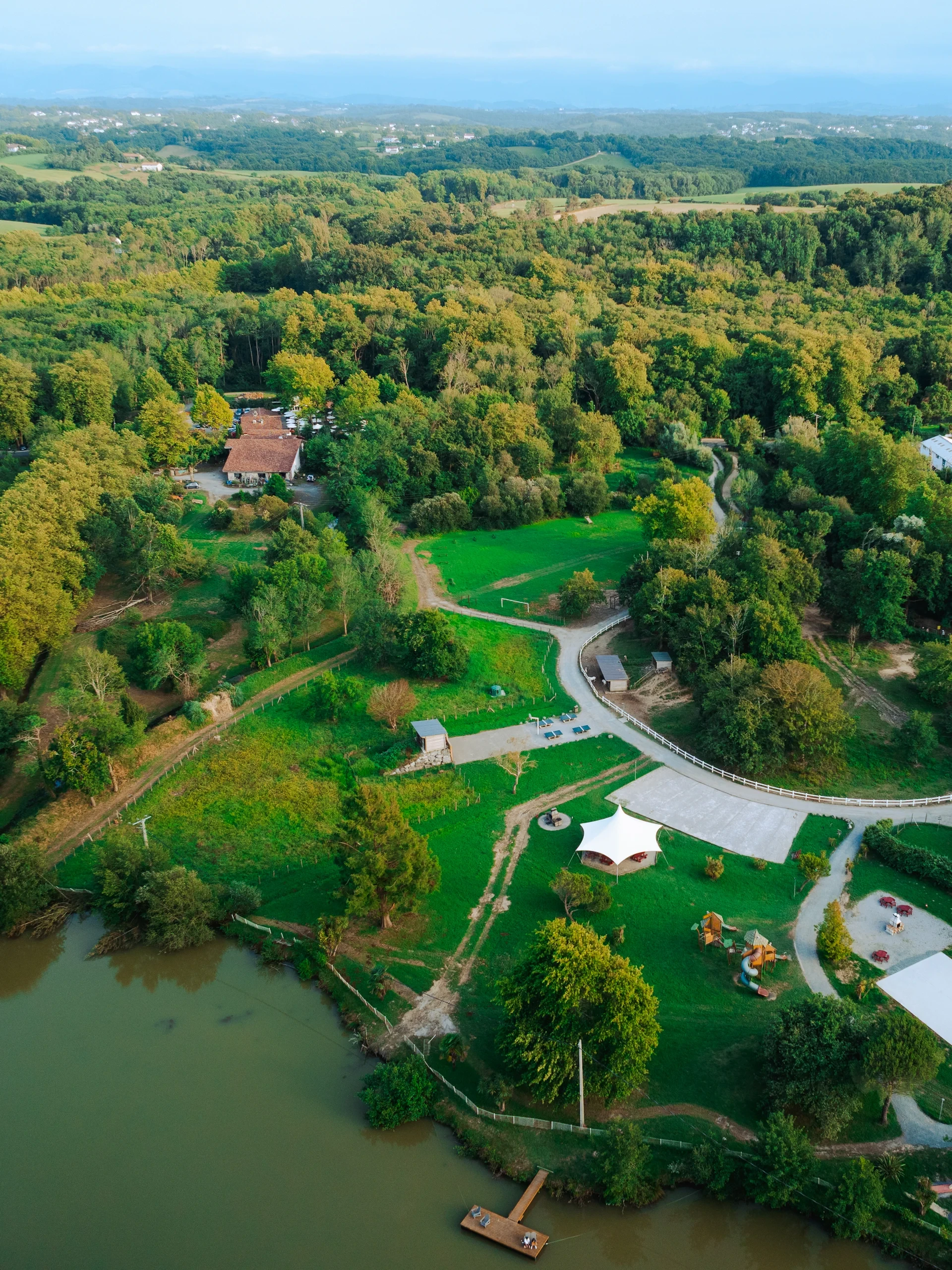 Vue aérienne du Camping le ruisseau, camping à Bidart, avec étang, ponton et espaces de jeux dans un environnement boisé.