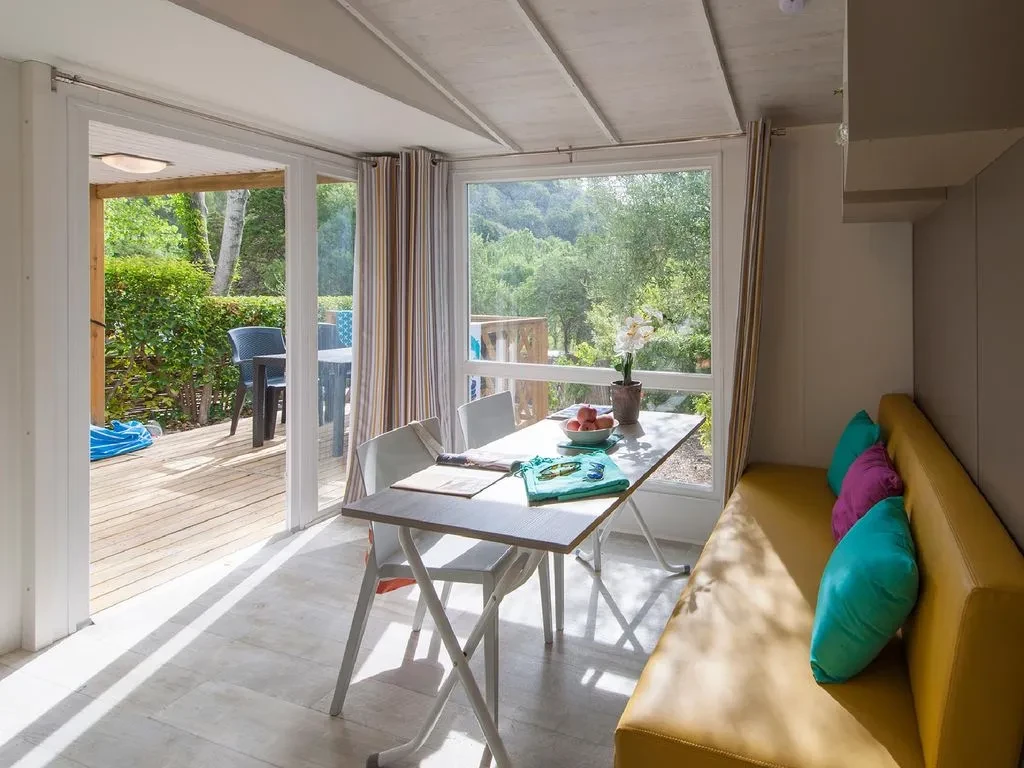 Séjour avec banquette jaune et table à manger devant de grandes baies vitrées ouvrant sur une terrasse en bois.