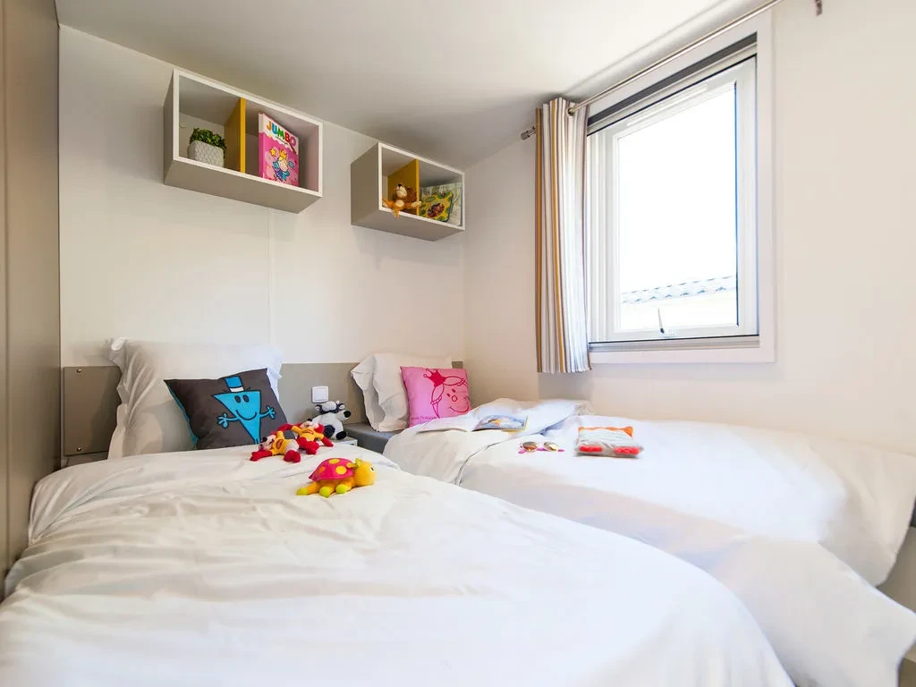 Chambre avec deux lits simples côte à côte, des coussins colorés et des peluches, éclairée par une fenêtre.
