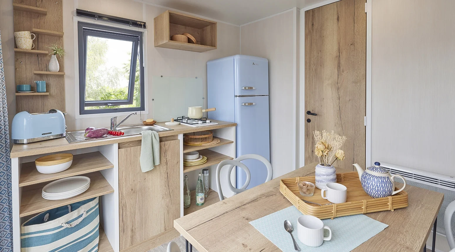 Cuisine avec réfrigérateur bleu clair, évier et plaque de cuisson, ouverte sur un coin repas avec table.