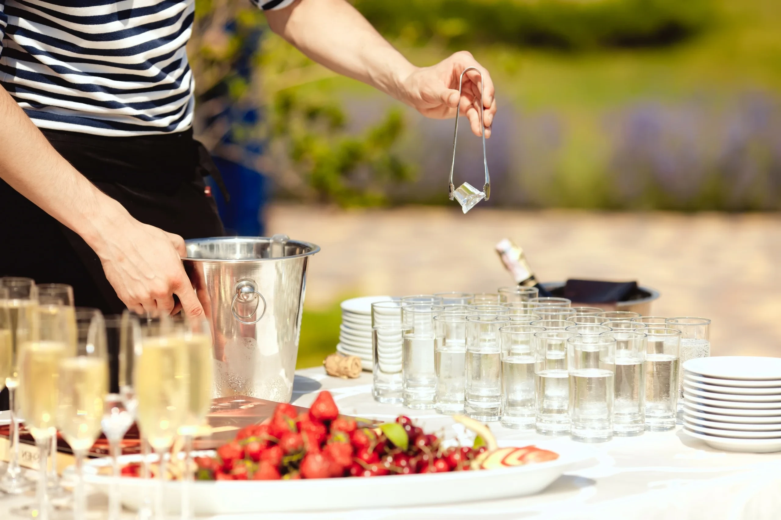 Une personne manipule un glaçon avec une pince au-dessus d’un buffet de réception en plein air.
