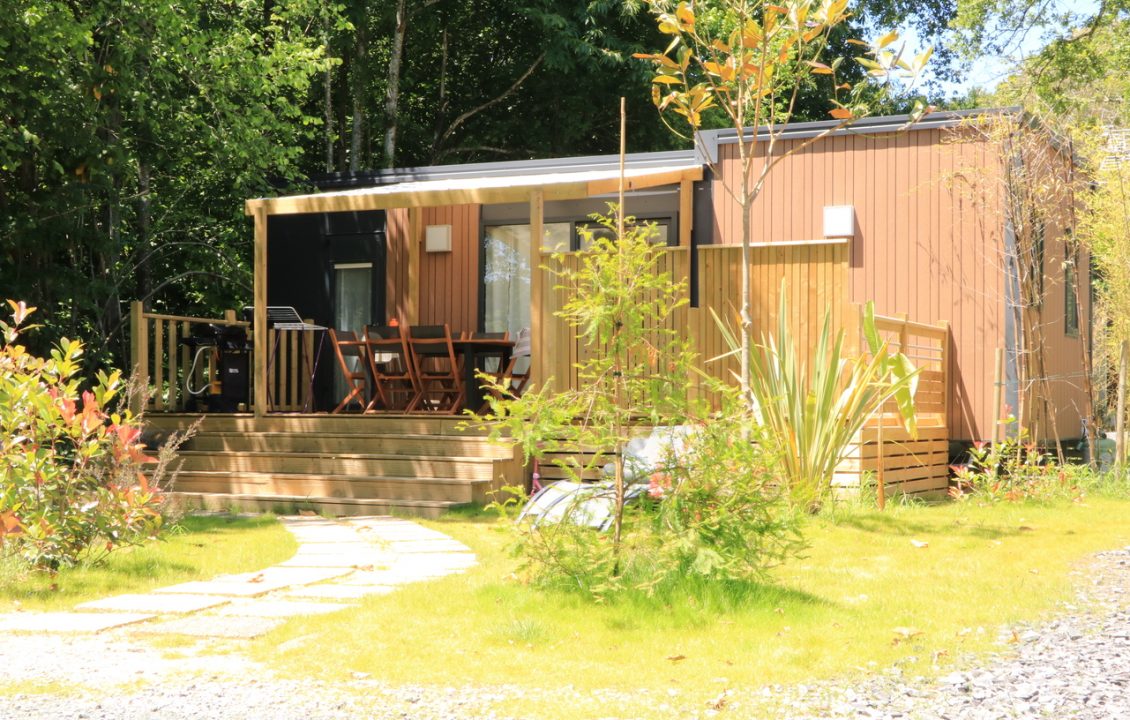 Cottage avec terrasse partiellement couverte et salon de jardin, entouré de végétation.