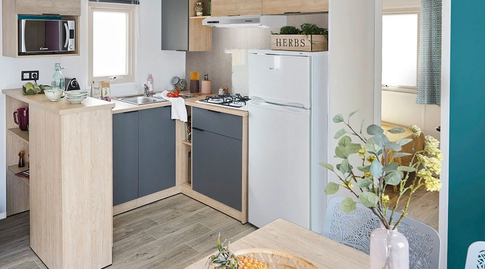 Cuisine en angle avec meubles en bois clair et gris, plaques de cuisson, réfrigérateur et micro-ondes.