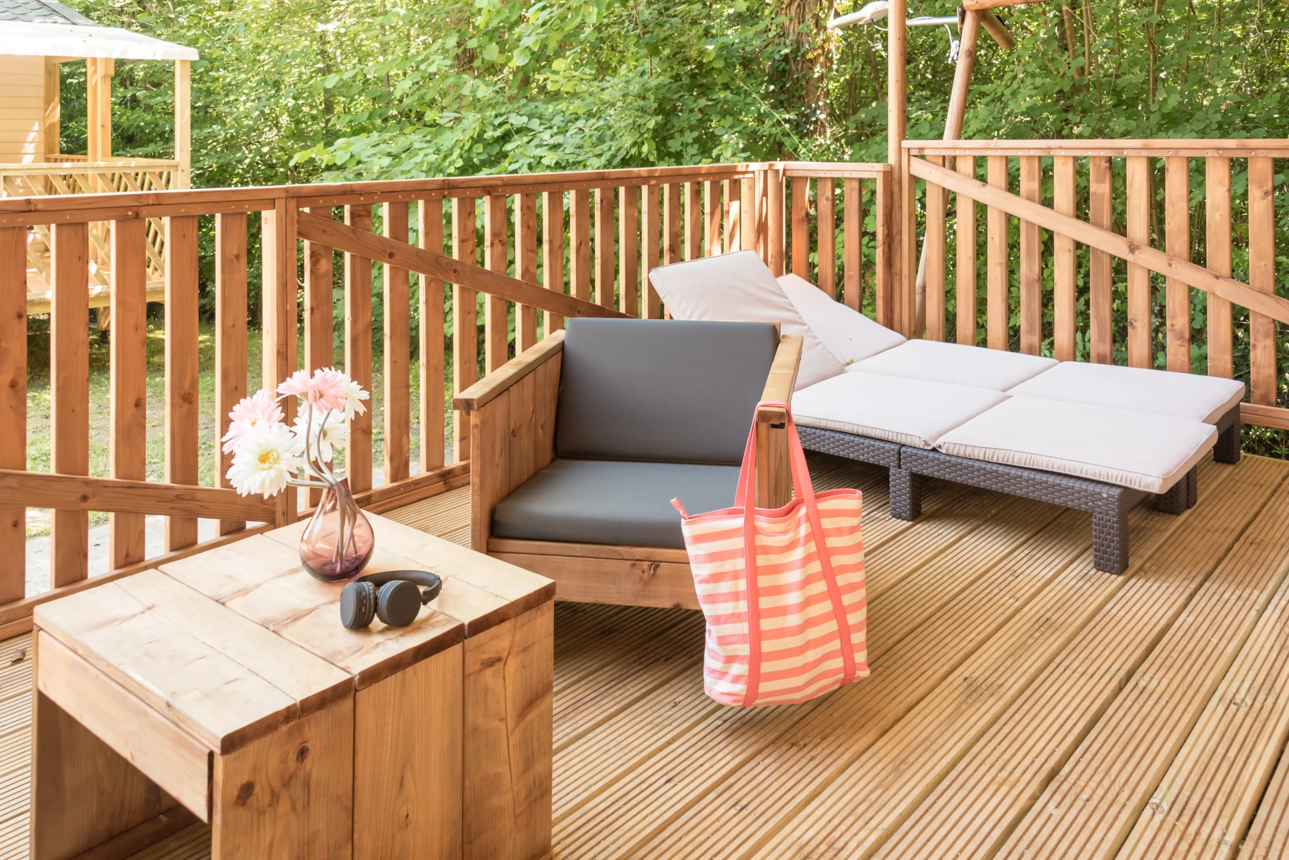 Terrasse en bois du Camping le ruisseau avec fauteuil, table basse et deux bains de soleil à coussins clairs.