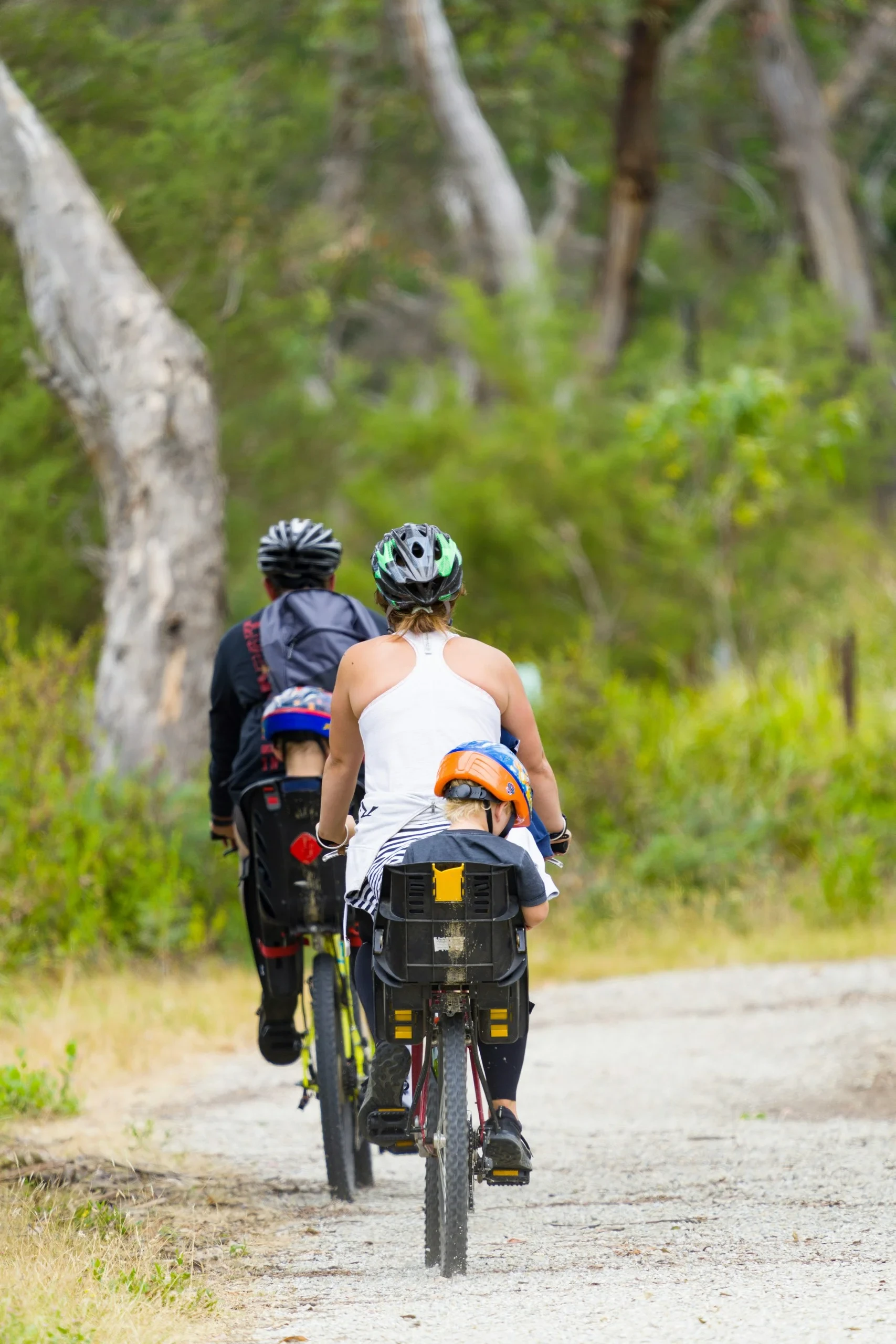 Deux adultes à vélo transportent chacun un enfant dans un siège arrière sur un chemin bordé d’arbres.