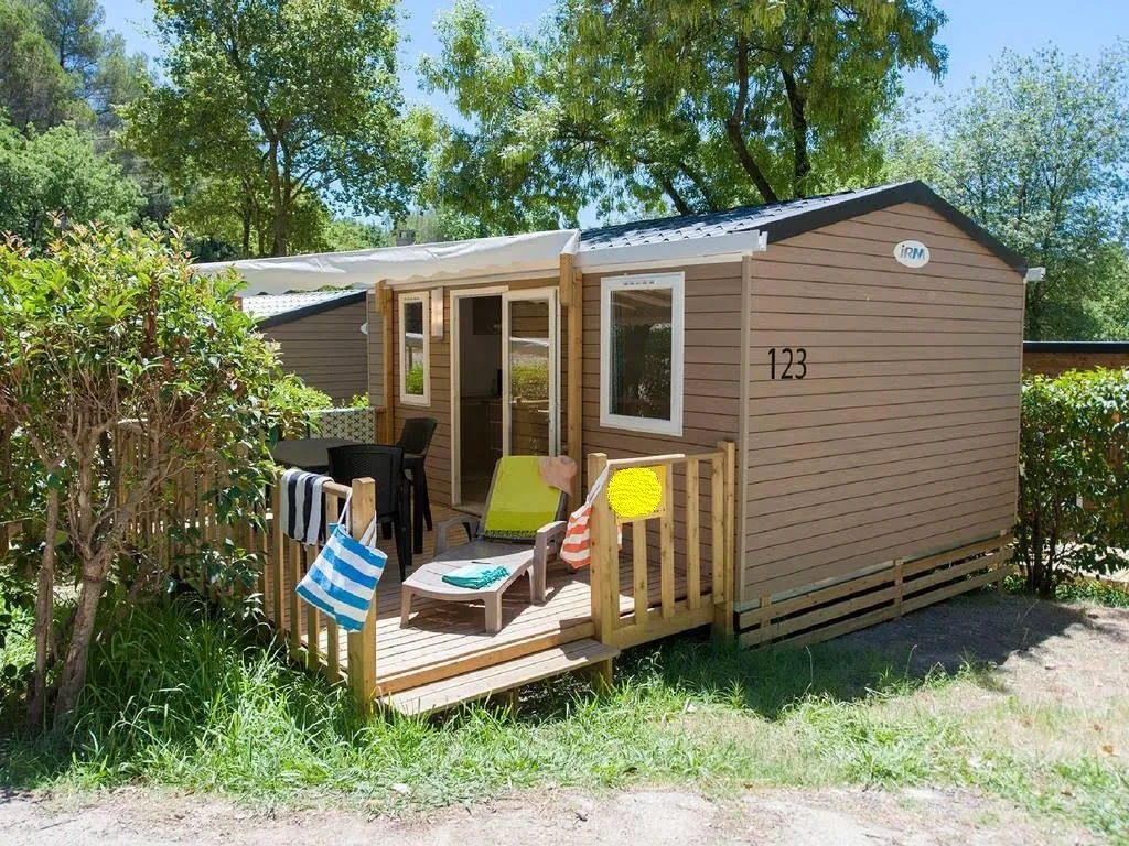 Mobil-home du Camping le ruisseau avec terrasse en bois, mobilier de jardin et transat, entouré de haies et d’arbres.