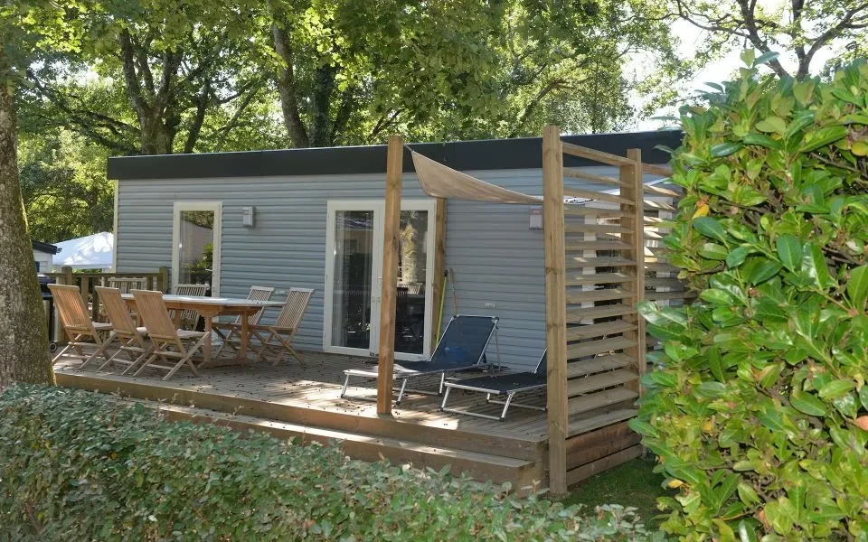 Mobil-home du Camping le ruisseau avec terrasse en bois, table de repas et deux transats sous les arbres.