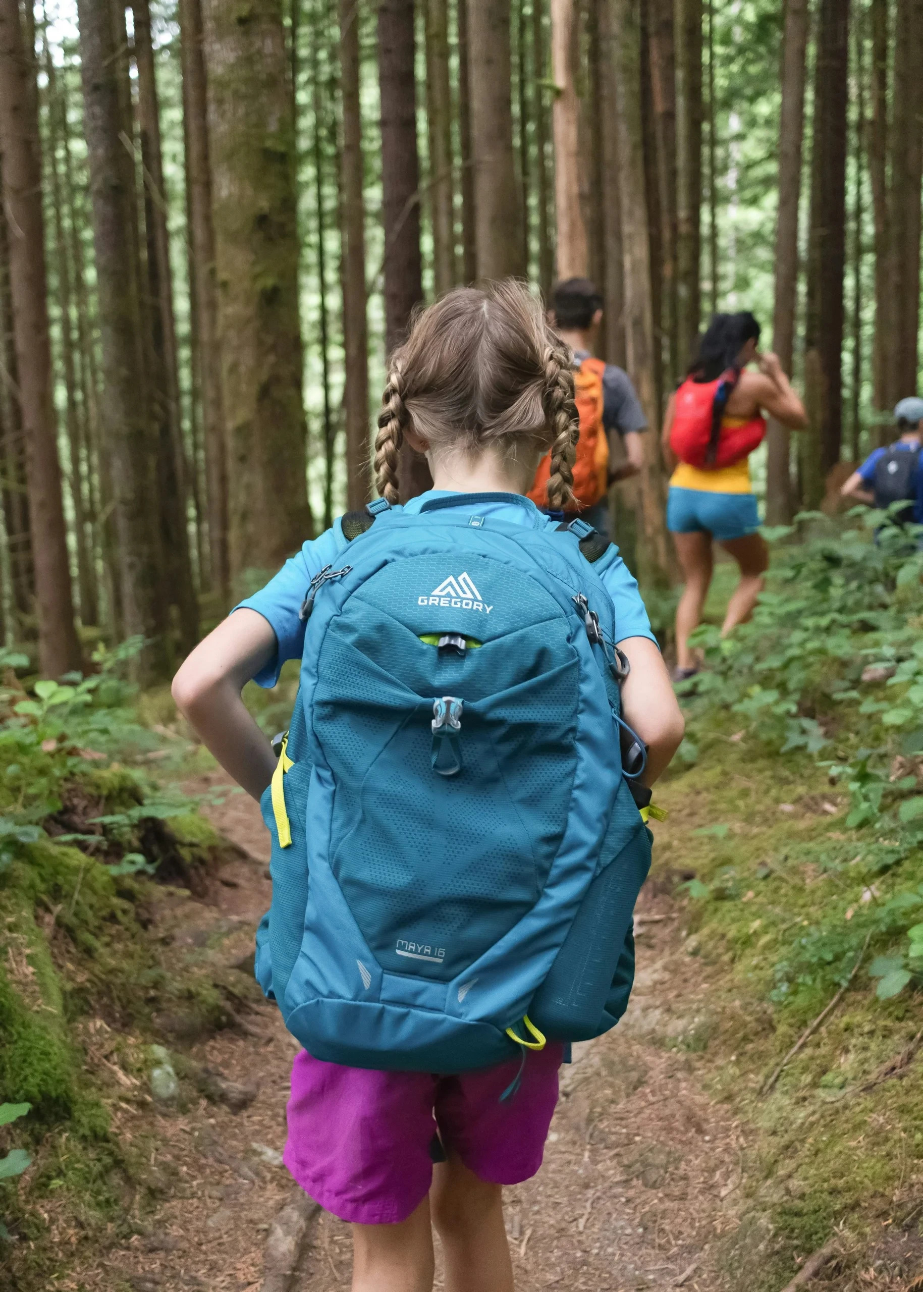 Enfant aux cheveux tressés portant un sac à dos bleu et suivant un groupe sur un sentier forestier.