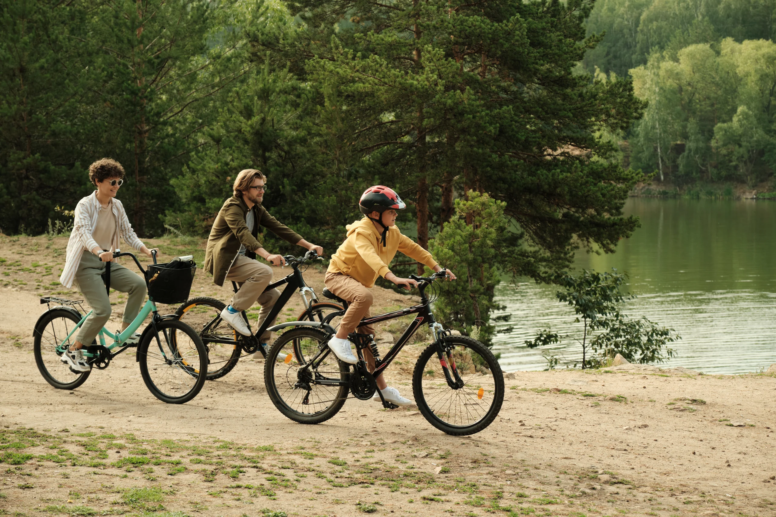 Deux adultes et un enfant casqué roulent à vélo sur un chemin au bord d’un lac arboré.