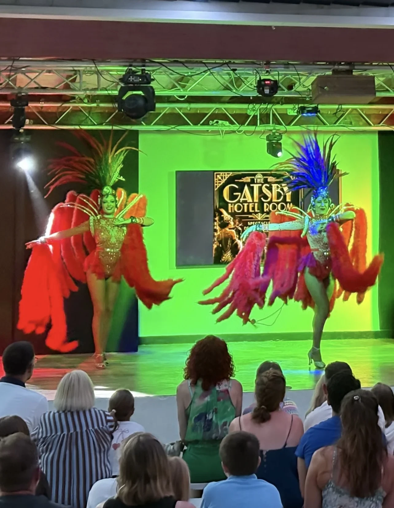 Deux artistes en costumes à plumes rouges et coiffes colorées se produisent sur scène devant un public assis.
