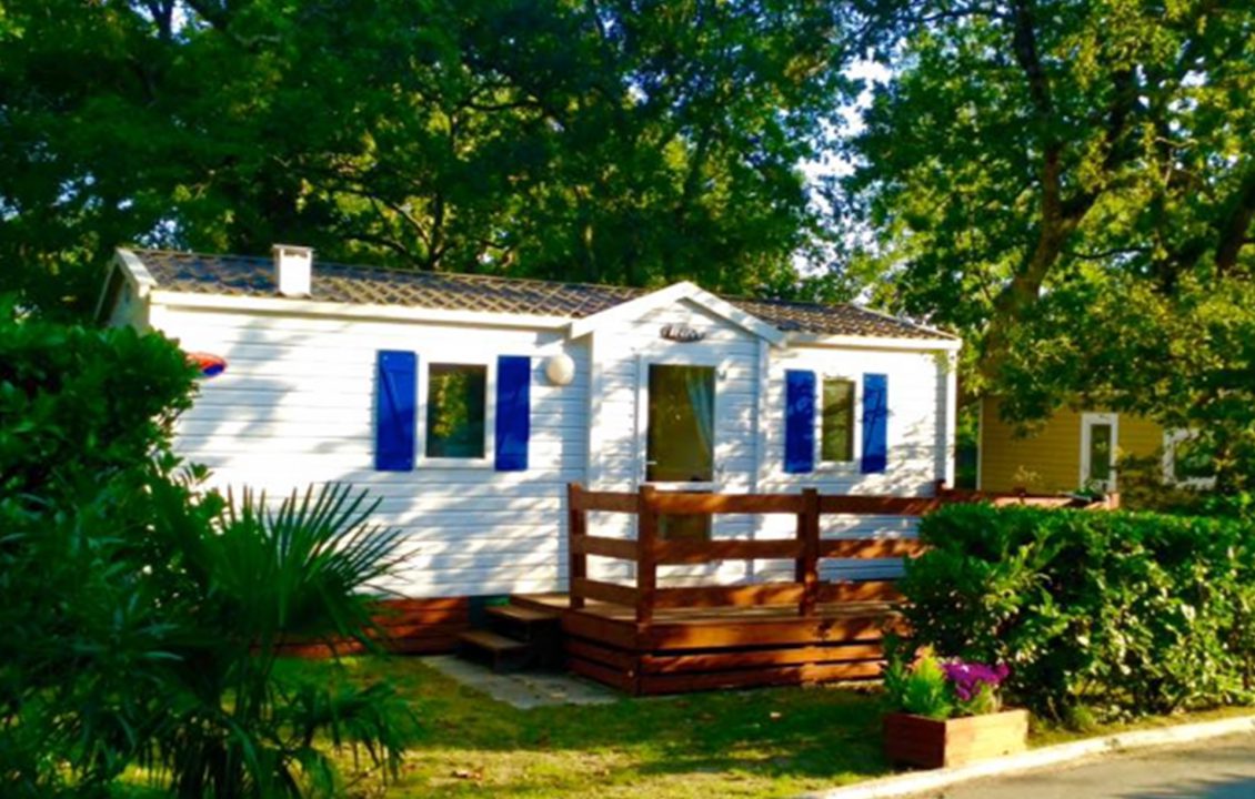 Mobil-home blanc aux volets bleus avec terrasse en bois arborée au Camping le ruisseau.