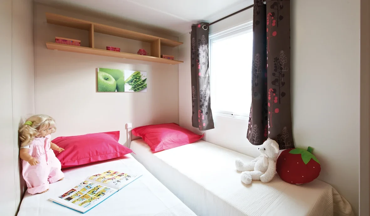 Chambre pour enfants avec deux lits simples côte à côte, des oreillers roses et une fenêtre bordée de rideaux à motifs.