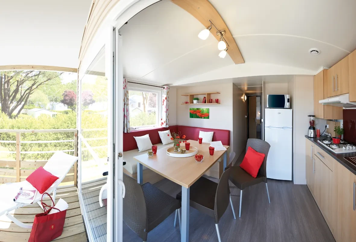 Séjour du Camping le ruisseau avec banquette d’angle, table à manger et cuisine ouverte, donnant sur une terrasse par une baie vitrée.