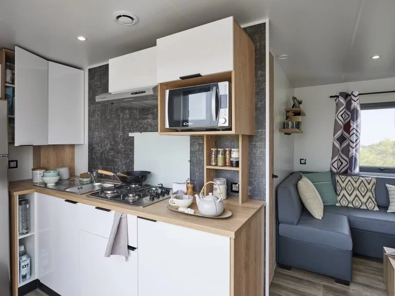 Cuisine ouverte sur le salon, avec plaque de cuisson au gaz, micro-ondes et rangements blancs et bois.