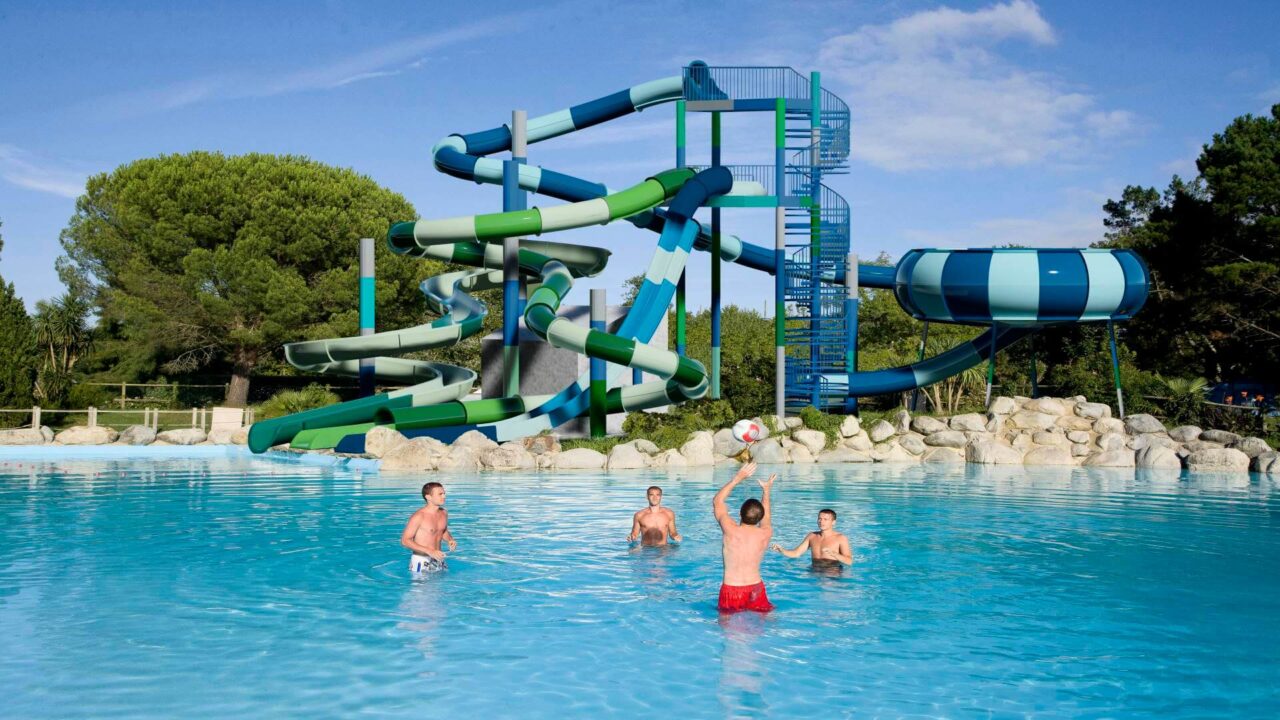 Baigneurs jouant au ballon dans une piscine devant plusieurs toboggans aquatiques.