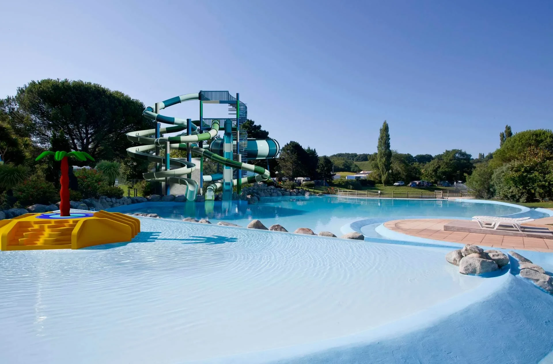 Bassins et toboggans du Camping le ruisseau, avec un jeu en forme de palmier dans le bassin peu profond et des transats au bord de l’eau.