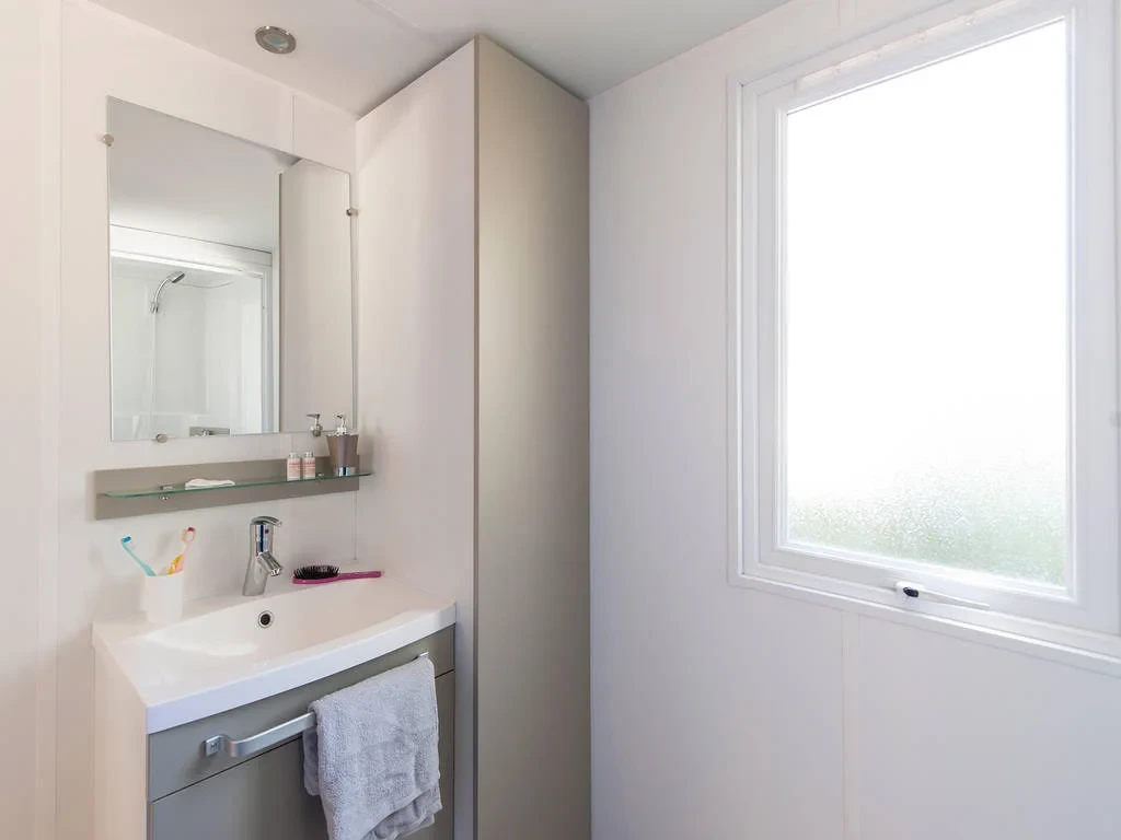 Salle d’eau avec meuble vasque, miroir et colonne de rangement à côté d’une fenêtre à vitrage dépoli.