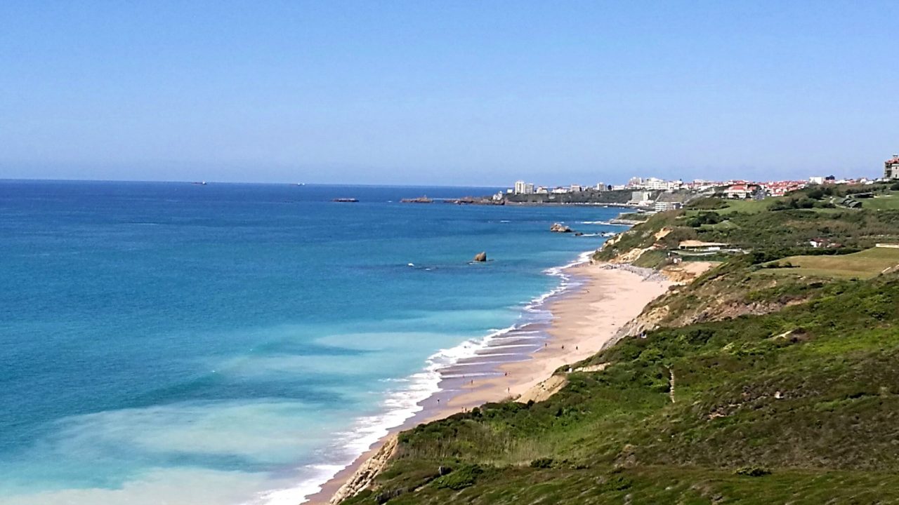 Plage de sable bordée de falaises végétalisées, avec des bâtiments au loin sur la côte.