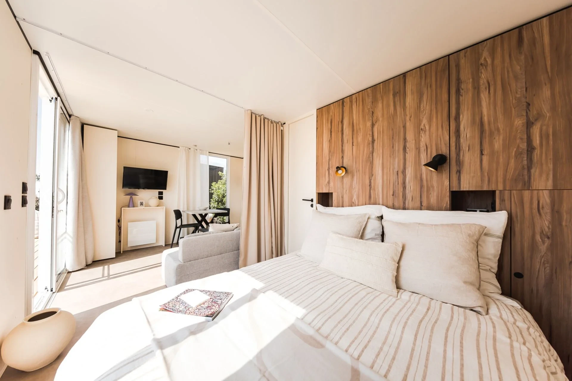 Lit double au premier plan et espace séjour avec canapé, table et télévision près des baies vitrées.