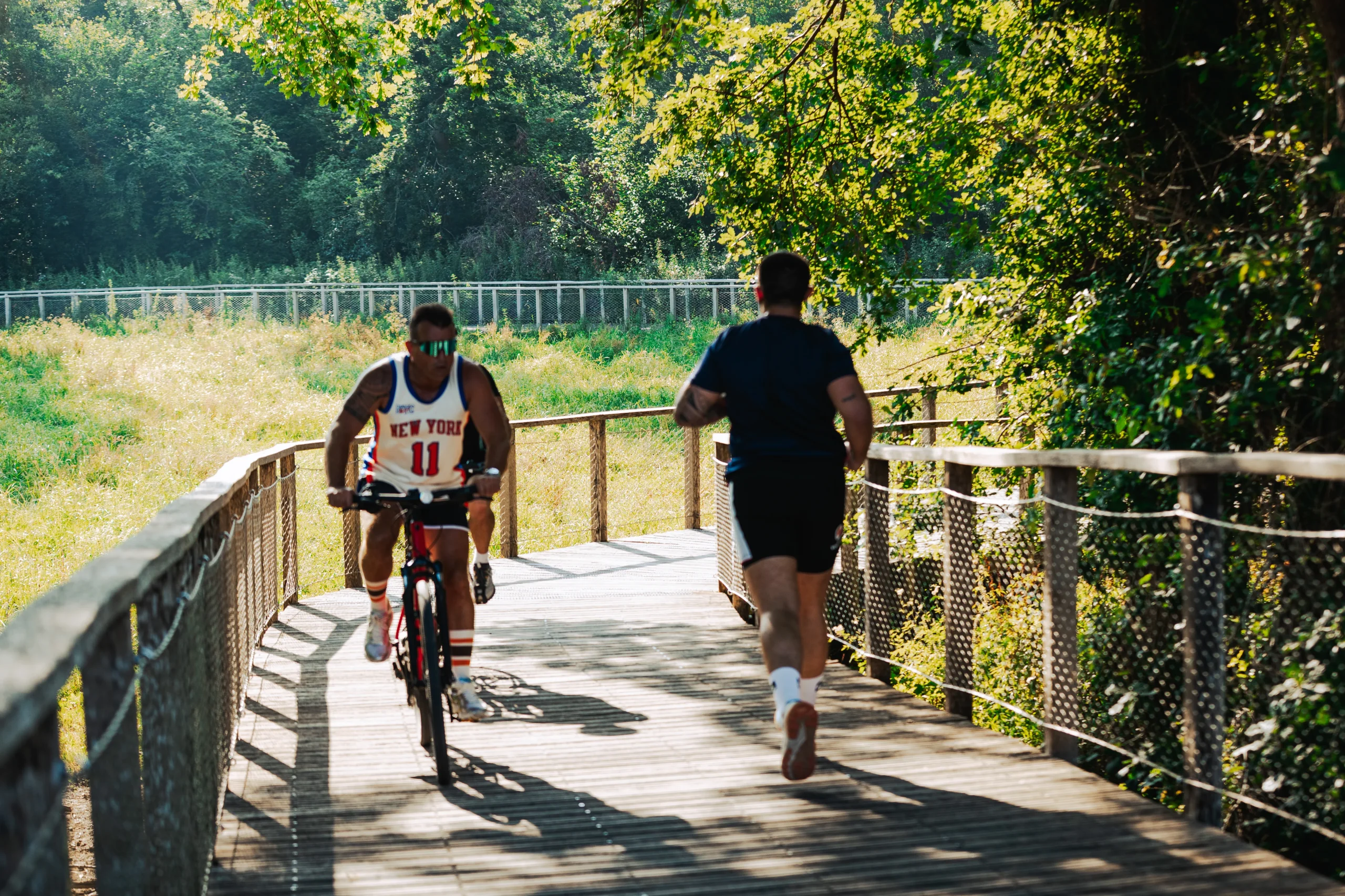 Un cycliste et un joggeur se croisent sur une passerelle en bois bordée de végétation.