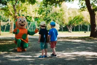 Deux enfants face à une mascotte orange et verte qui leur ouvre les bras dans un espace arboré.