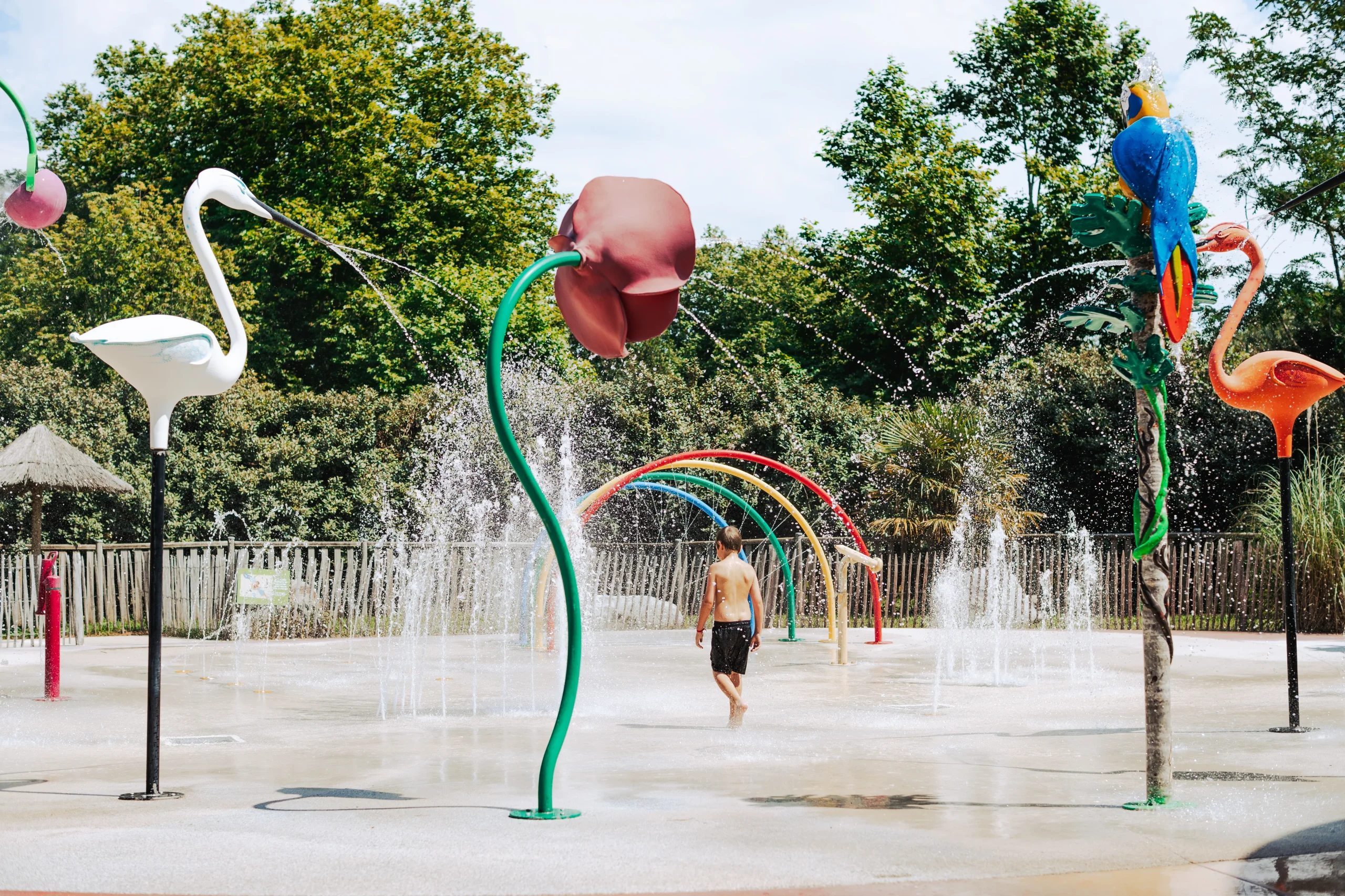 Un enfant traverse les jets d’eau du Camping le ruisseau, camping en bord de mer à Bidart, entre des arches colorées et des sculptures d’oiseaux.