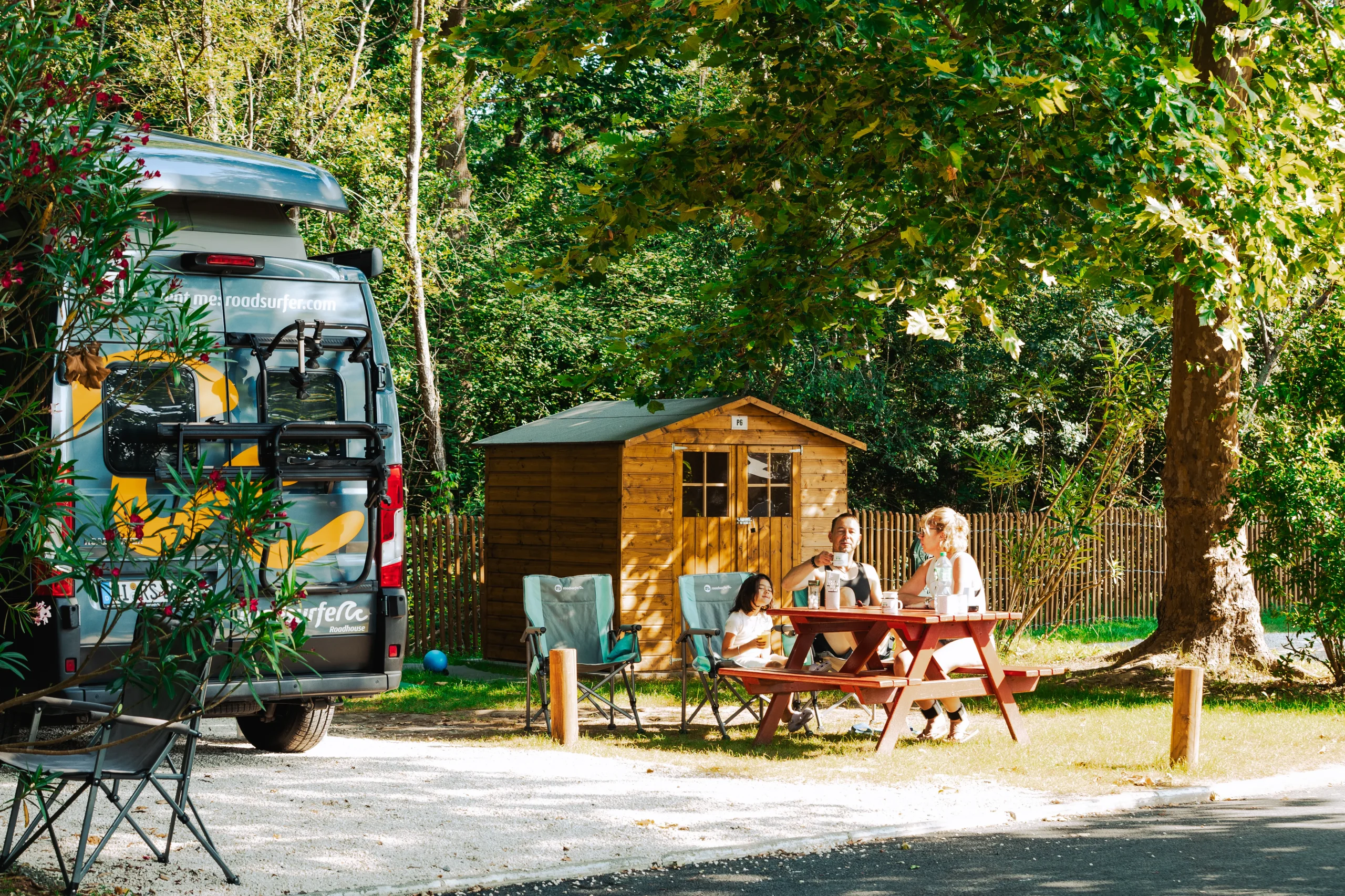 Des vacanciers attablés sur une parcelle arborée, à côté d’un van aménagé et d’un cabanon en bois.