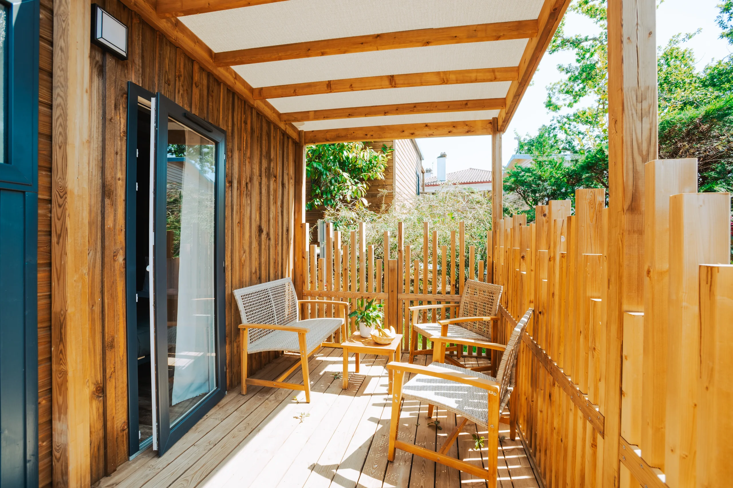 Terrasse couverte en bois avec une banquette, deux fauteuils et une petite table, bordée de palissades ajourées.