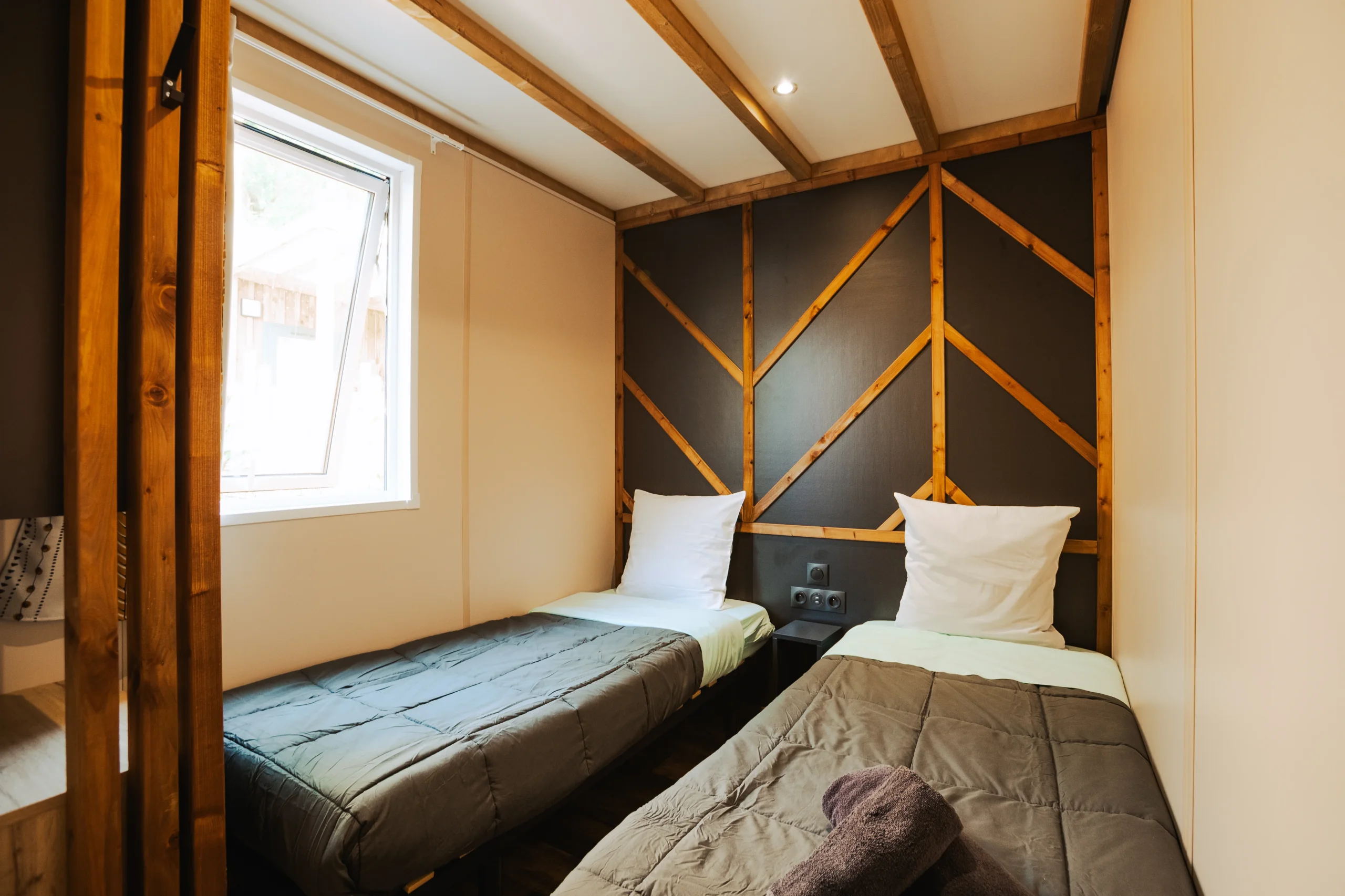 Chambre à deux lits simples séparés par une petite table de chevet, avec poutres apparentes et fenêtre latérale.