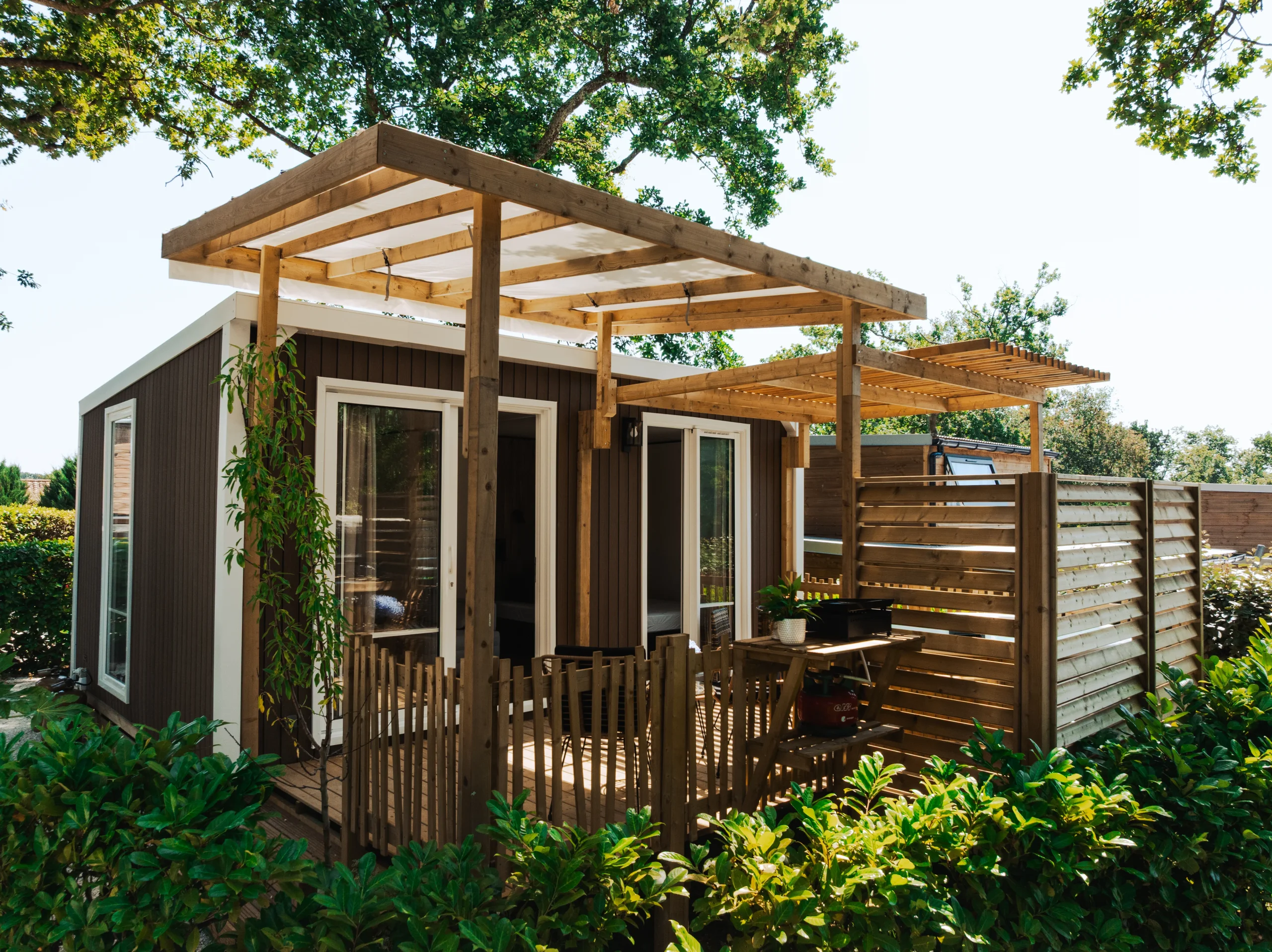 Hébergement du Camping le ruisseau avec grandes baies vitrées et terrasse en bois sous pergola, entouré de végétation.