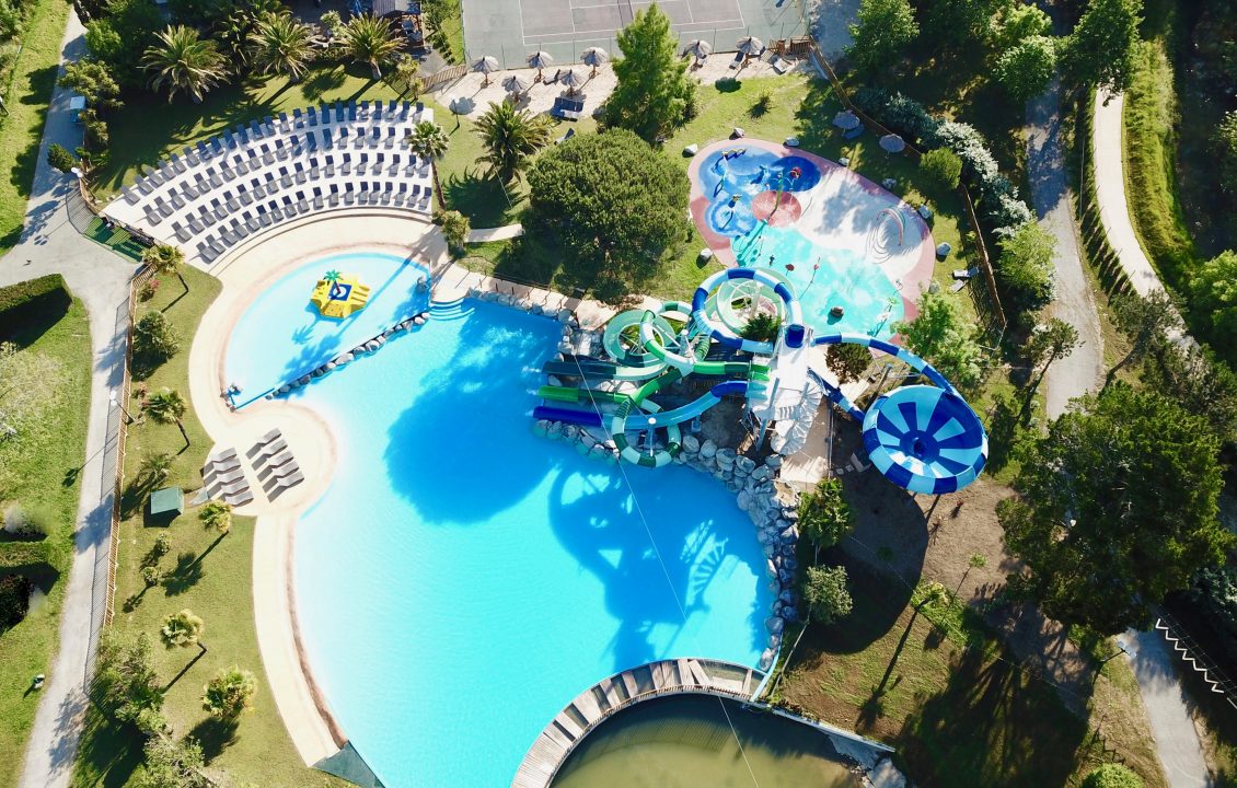 Vue aérienne élargie du parc aquatique du Camping le ruisseau, entouré de plages avec transats et d’espaces arborés.