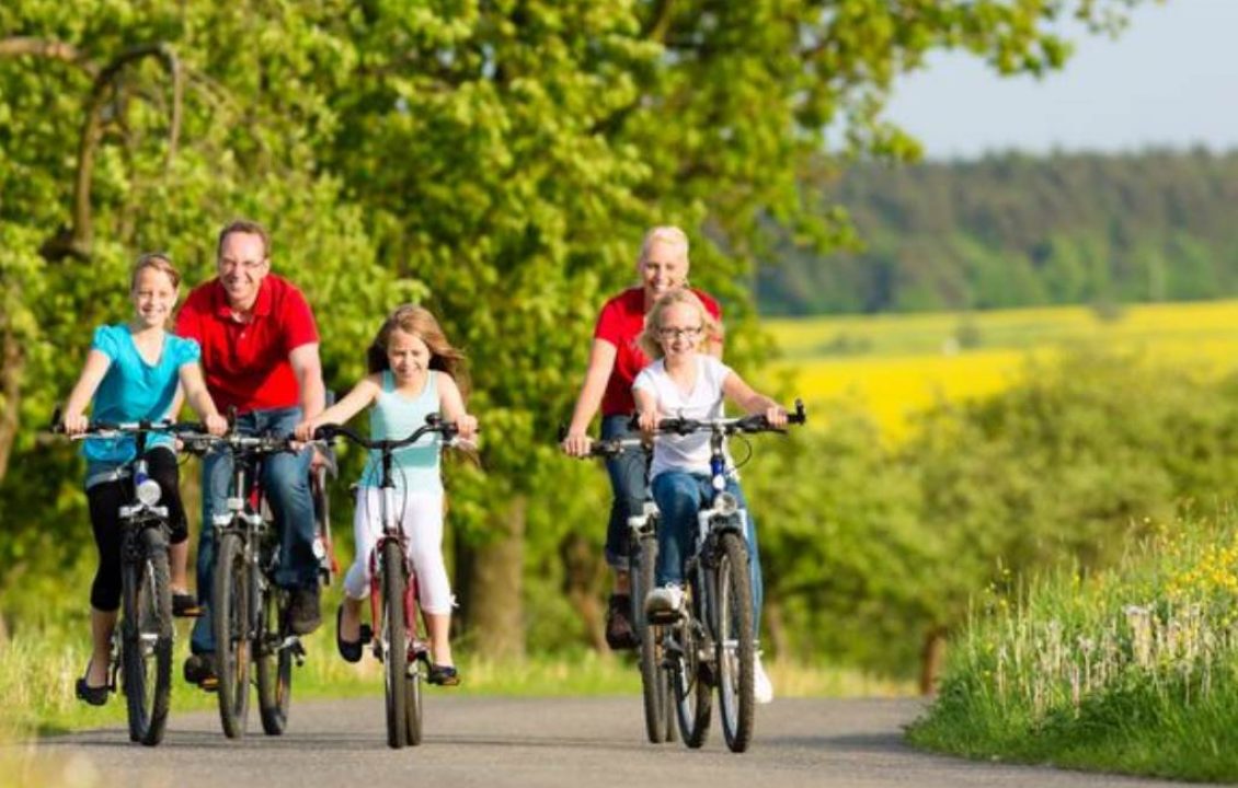 Adultes et enfants à vélo sur un chemin bordé d’arbres et de champs fleuris.