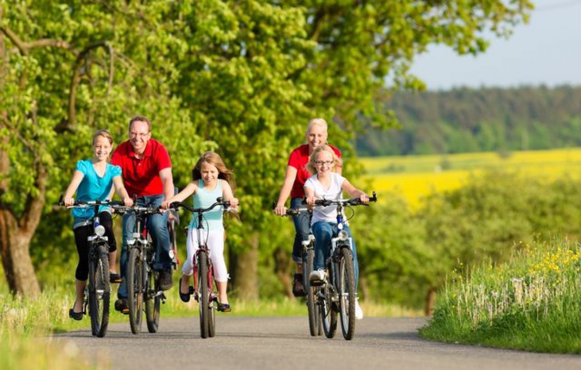 Deux adultes et trois enfants à vélo sur un chemin bordé d’arbres et de champs.